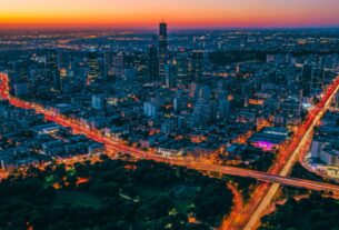 Vista aérea de cidade ao entardecer com ruas iluminadas formando uma rede de luz entre áreas verdes e edifícios