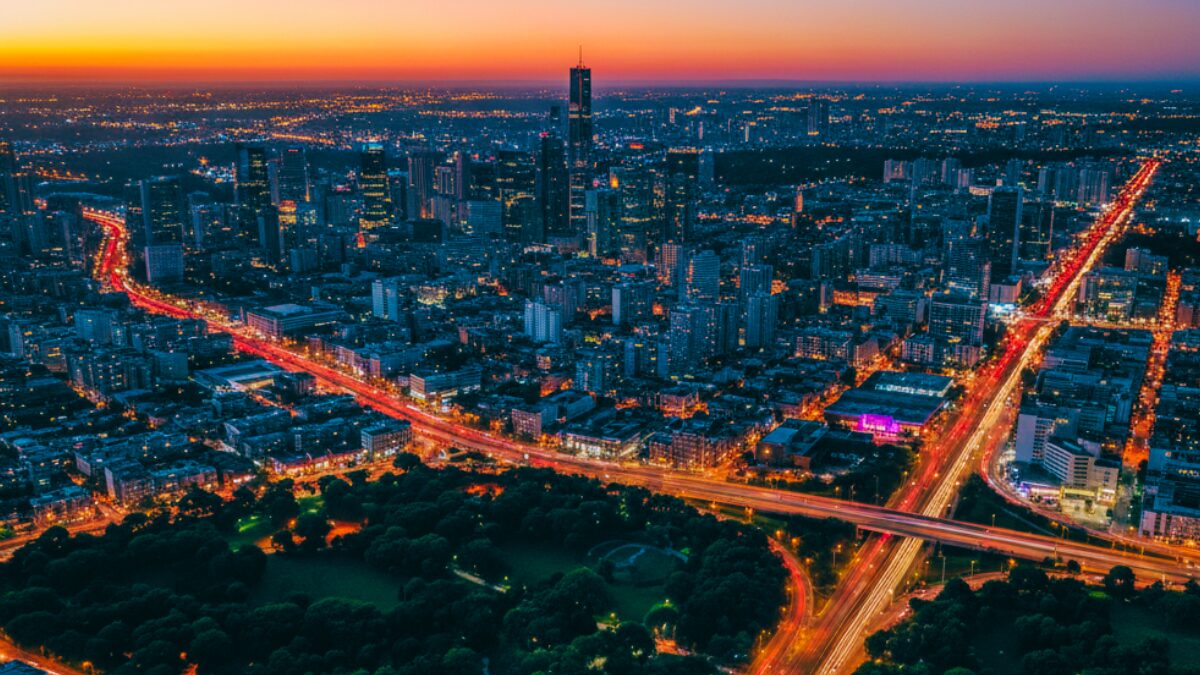 Vista aérea de cidade ao entardecer com ruas iluminadas formando uma rede de luz entre áreas verdes e edifícios